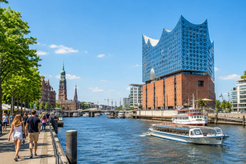 Solrig udsigt over Hamborgs havneområde med Elbphilharmonie, både på vandet, hyggelig promenade og mennesker langs kanalen på en varm sommerdag.